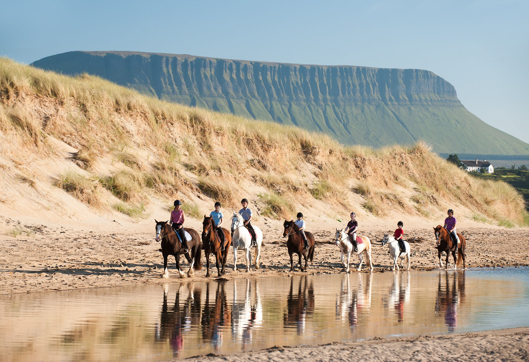 Des cavaliers progressent sur une plage irlandaise, une montagne abrupte se dessine à l'horizon sous un ciel dégagé.