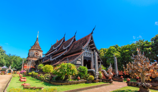 Temple traditionnel thaïlandais avec toit en teck sculpté, jardins luxuriants et ciel bleu clair.