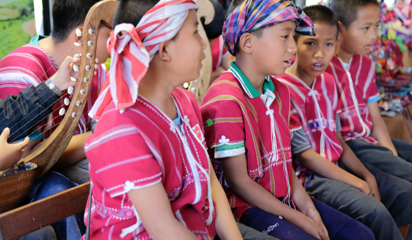 Enfants en vêtements traditionnels thaïlandais rouges, assis ensemble, l'un joue de la harpe, lors d'un événement culturel.