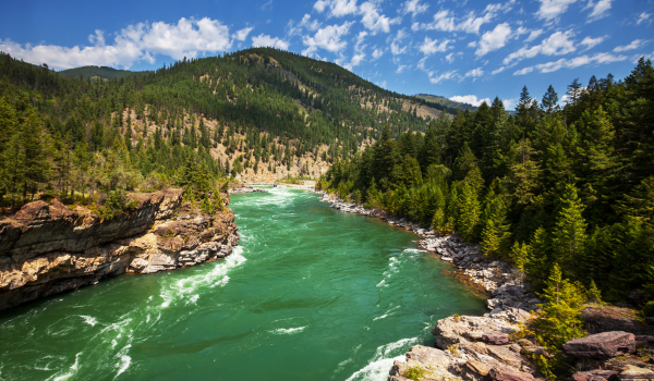 Rivière turquoise serpentant à travers une vallée boisée avec des montagnes verdoyantes sous un ciel partiellement nuageux.
