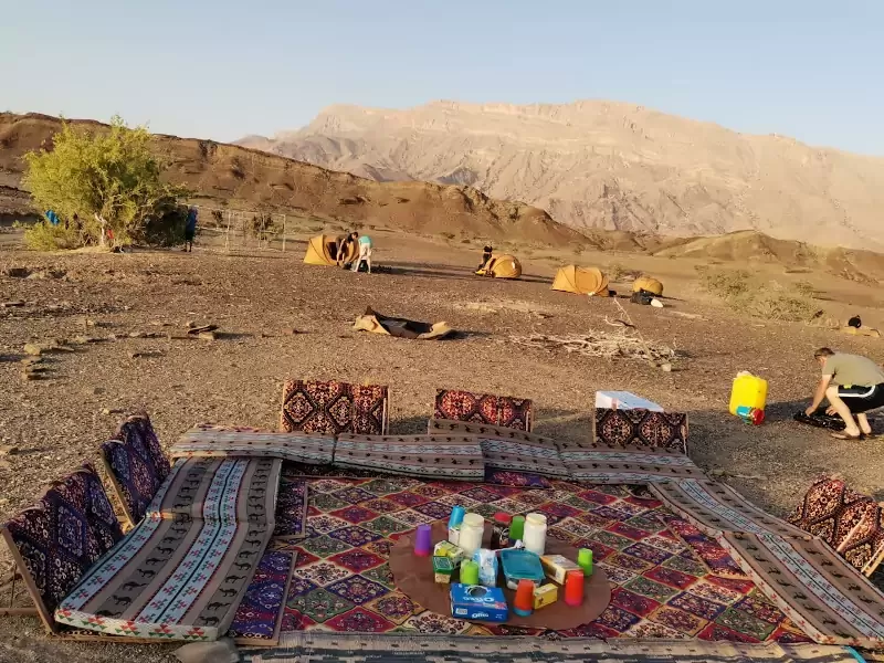 Espace de bivouac dans le désert avec tapis et coussins colorés, montagnes en arrière-plan, tentes installées autour.