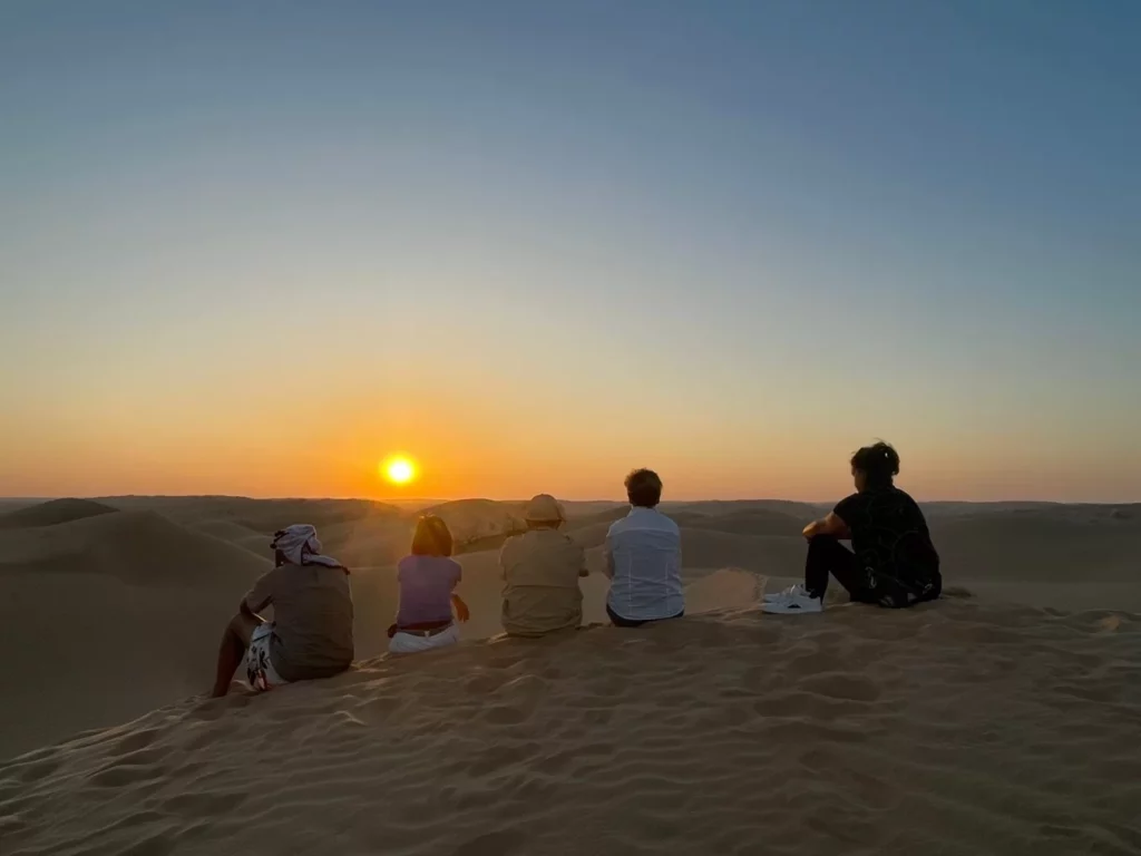 Cinq personnes assises sur une dune de sable au coucher du soleil, contemplant l'étendue désertique.