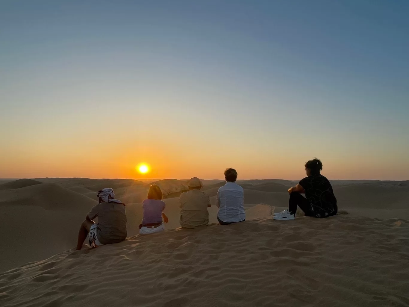 Cinq personnes assises sur une dune de sable au coucher du soleil, contemplant l'étendue désertique.