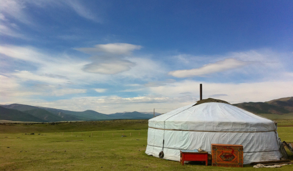 Yourte mongole sur une prairie verdoyante sous un ciel bleu, montagnes à l’horizon.