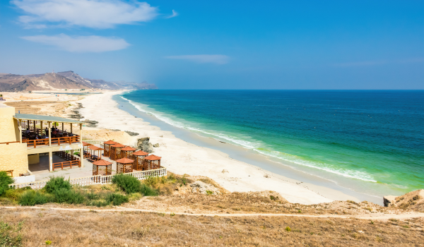 Plage de sable blanc bordée par une mer turquoise, vue sur un hôtel avec terrasses et collines en arrière-plan.