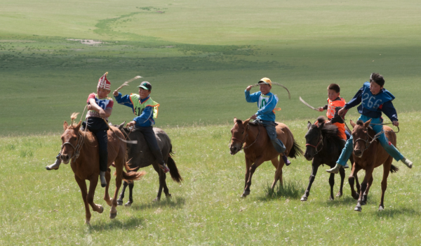 Cinq cavaliers en tenue colorée galopent sur une plaine verte, chacun tenant un bâton pendant une course.